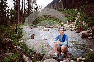 Turistka Sedin na kameni pri horskej rieke vo Vysokých Tatrách na Slovensku. Oblečená v modrom tričku, šedých šortkách, Turistika