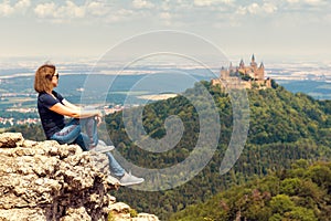 Girl tourist sitting on cliff looks at Hohenzollern Castle on mountain top, Germany