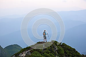 The girl with the touristic equipment goes up to the peak of the rocky high hill with the lawn. The scenery of the mountains.