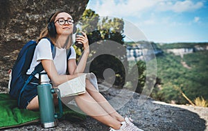 Girl traveler closed her eyes with pleasure enjoying music in headphones and drinking tea from thermos sitting in mountains