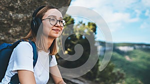 Girl traveler enjoys music with headphones dreaming in mountains in the fresh cleare air, young female tourist with glasses