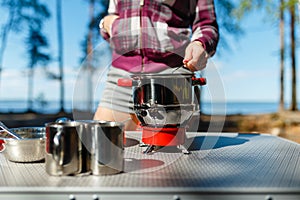 Girl traveler prepares food on portable gas stove, on a folding table on the background of camping in forest. Women's hands inter