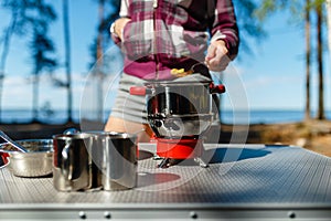 Girl traveler prepares food on portable gas stove, on a folding table on the background of camping in forest. Women's hands inter