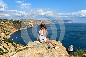 The girl traveler sits on a huge stone on a rock and looks at the sea