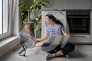 Girl turned powerful electric fan enjoying cool wind sitting on floor home work with laptop.