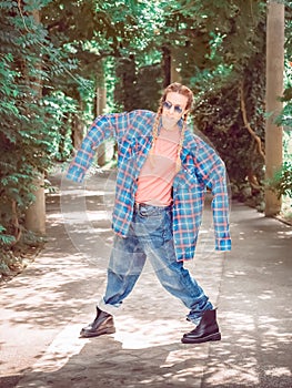 A girl with two pigtails in a plaid shirt with oversleeves and oversized jeans stands in a pose on a green alley. Dance