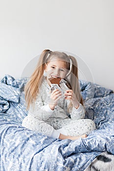 A girl with two ponytails in soft gray pajamas sits on a bed and bites a bar of chocolate in foil