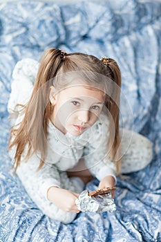 A girl with two ponytails in soft gray pajamas sits on a bed and bites a bar of chocolate in foil