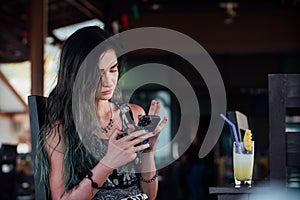 The girl typing in the phone, sitting in a cafe with fruit pineapple juice in a glass and a tube.