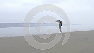 Girl with umbrella walking towards on sandy beach from left to right.