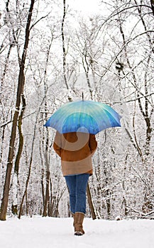 Girl under blue umbrella in snowy forest. Snowfall concept. Woman under wet snow rain in winter park.