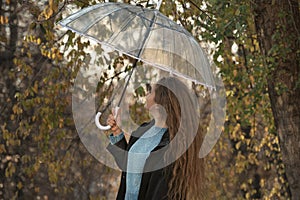 Girl under transparent umbrella in black cloak walk in park. Young woman with wavy long hair. Back view