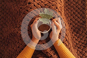 Girl under a warm blanket holding a white mug with a hot drink and a maple leaf in her hands