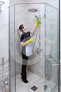 Girl in uniform cleaning the shower stall. Bathroom interior with marble tiles. The concept of cleanliness and hygiene.