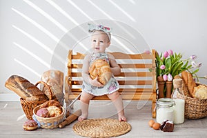 A girl up to a year old in a kitchen apron and a bow on her head sits on a bench, next to tartlets with booths