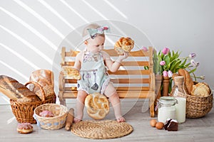 A girl up to a year old in a kitchen apron and a bow on her head sits on a bench, next to tartlets with booths