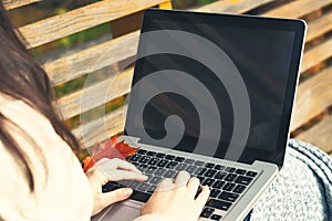 Girl uses a laptop while sitting on a bench in the autumn park. Blank for design on laptop screen