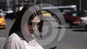 A girl uses a mobile phone on the street on a sunny day against the background of a passing car stream. The girl uses