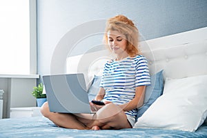 Girl using laptop and smartphone while sitting on bed. Woman texting sms, working on laptop online