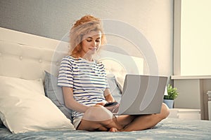 Girl using laptop and smartphone while sitting on bed. Woman texting sms, working on laptop online