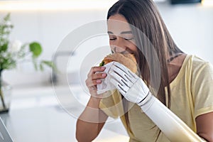 Girl using prosthetic arm while eating at home, woman with disability holds croissant in artificial prosthetic limb