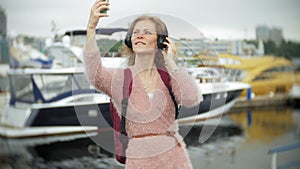 A girl using a smartphone on the beach, listening to music in headphones, dancing a yacht and sailing in the harbor.