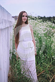 A girl in a white dress is standing in a field by the fence