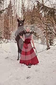 A girl in a vintage red dress stands next to a horse in a winter forest.