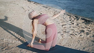 Girl in violet tracksuit practises camel yoga pose on sand