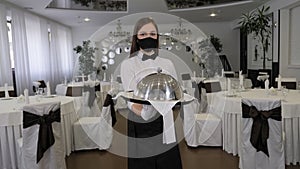 A girl waitress in a mask in a restaurant, she holds a tray with a dish.