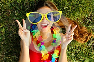 Girl wake up on grass after Carnival party. Young woman with big funny sunglasses and carnival garland lying on grass showing