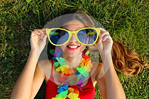 Girl wake up on grass after Carnival party. Young woman with carnival garland holds big funny sunglasses lying on grass