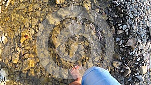 Girl walking barefoot on stones of shallow river close-up, sunny summer day