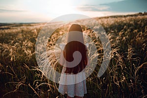 Girl walking on a wheat field holding wheat spike at beautiful sunset. Freedom and fresh air concept.