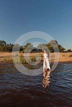A girl walks in the water at dusk holding a wet white summer dress