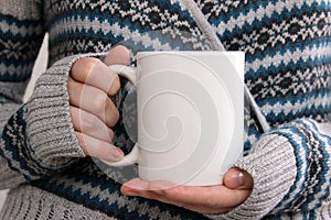 Girl in a warm cardigan is holding white mug in hands.