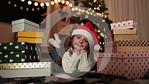 Girl in warm knitted white sweaters and red santa hat lying on floor in front of Christmas tree