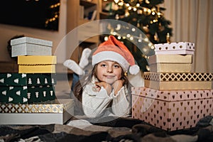 Girl in warm knitted white sweaters and red santa hat lying on floor in front of Christmas tree