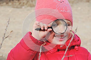 A girl in warm red clothes with a magnifying glass in her hand examines a tree branch .  Winter outdoor kids activity concept