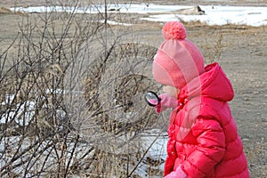 Girl in warm red clothes with a magnifying glass in her hand examines a tree branch .  Winter outdoor kids activity concept