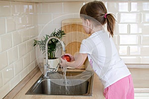 Girl washes a red apple in the kitchen near the sink. The concept of children`s health and independence