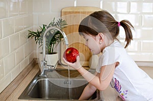 Girl washes a red apple in the kitchen near the sink. The concept of children`s health and independence