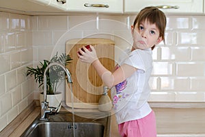 Girl washes a red apple in the kitchen near the sink. The concept of children`s health and independence