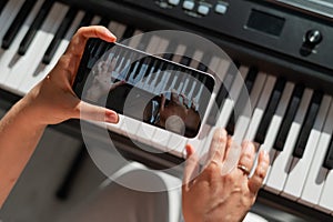 A girl watches an online music lesson on a smartphone and learns to play the piano. Close-up of woman`s hands on keys.