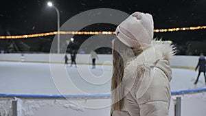 A girl watches people on a skating rink in the city.