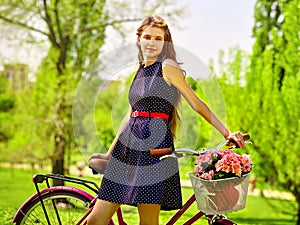 Girl wearing blue polka dots sundress rides bicycle with flowers basket.