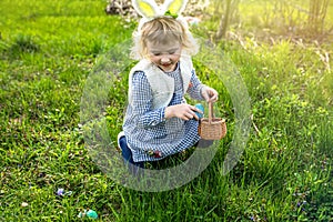 girl wearing bunny ears playing egg hunt on Easter. A child celebrates Easter. Girl sitting on grass with a basket full