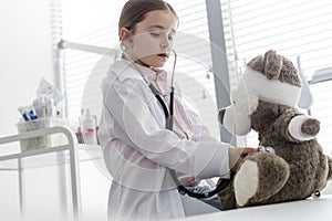 Girl wearing labcoat imitating doctor while examining teddybear with stethoscope at hospital