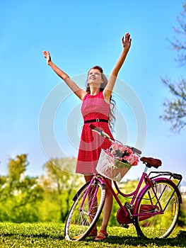 Girl wearing red dress hand up near bicycle with flowers .