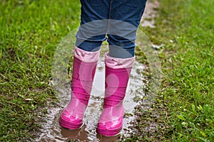 Child in bright pink rubber boots and blue jeans frolic in a dirty puddle